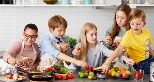 Fem barn i ett kök sträcker sig efter olika matvaror på ett bord.